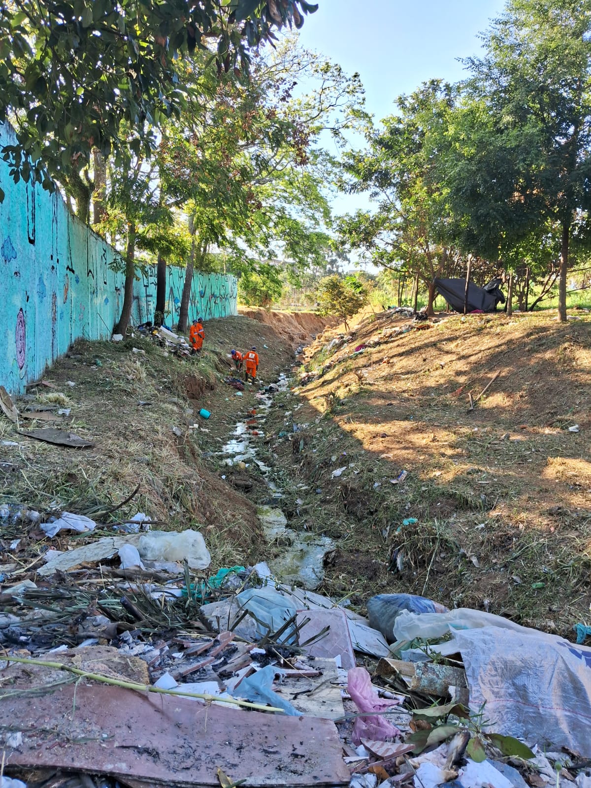 Córrego passando, lixo espalhado nas laterais e na parte de terra. Algumas pessoas em uniforme laranja limpando o local. 